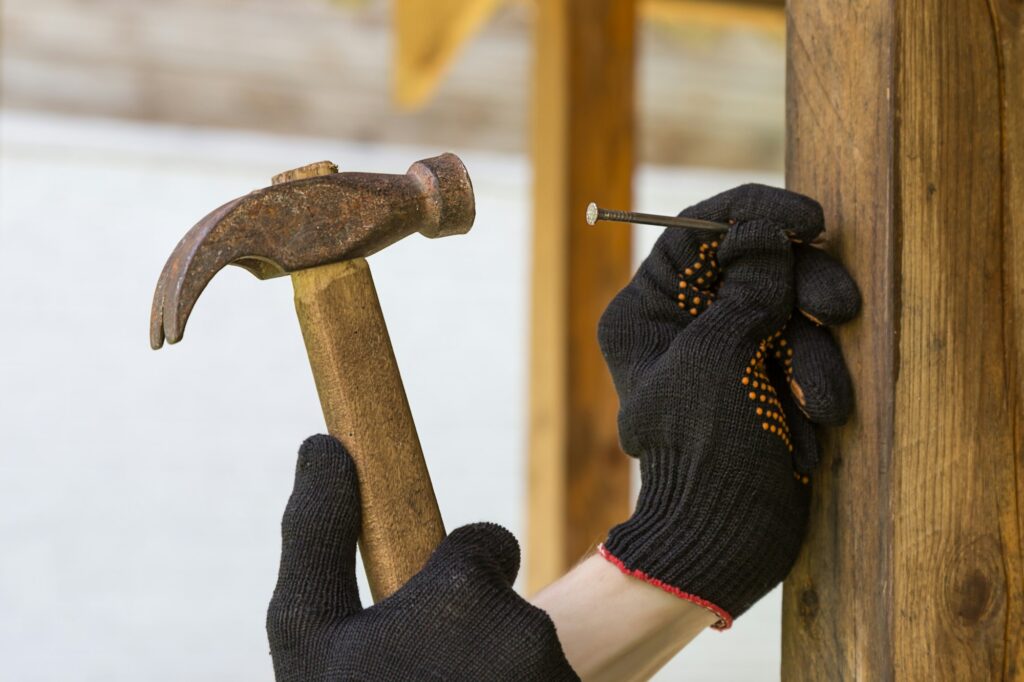 Hand in a construction glove hammers a nail into a wooden beam with a hammer